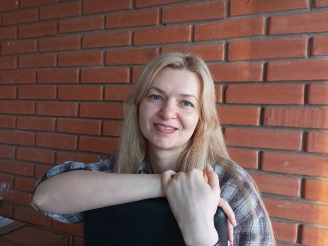 MelissaTyler with blonde hair, smiling, holding a black object, wearing a plaid shirt, arms crossed, brick wall background, blue eyes, fair skin, slight pimple on her cheek.