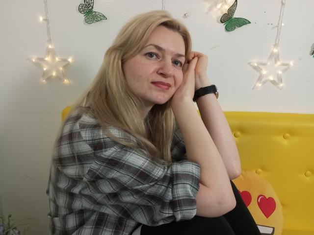 MelissaTyler with blonde hair, wearing a plaid shirt, black pants, and a black wristwatch, sitting on a yellow couch, resting her chin on her hand, smiling subtly.