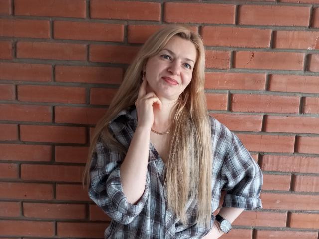 MelissaTyler with long blonde hair, wearing a plaid shirt, smiling slightly, hand on cheek, red brick wall background, wristwatch on left wrist.