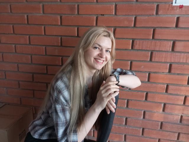 MelissaTyler with long blonde hair, smiling, wearing a plaid shirt, leaning on a black chair, showing her cute tits and a wristwatch, against a red brick wall.