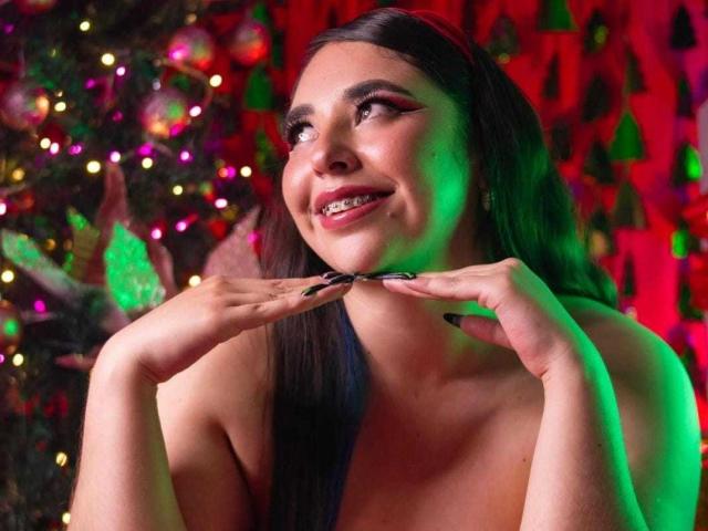 BaGiolett con lunghi capelli neri, sorridente, che si tocca il mento, con luci verdi e rosse sul viso, albero di Natale colorato con luci sullo sfondo, spalle scoperte..