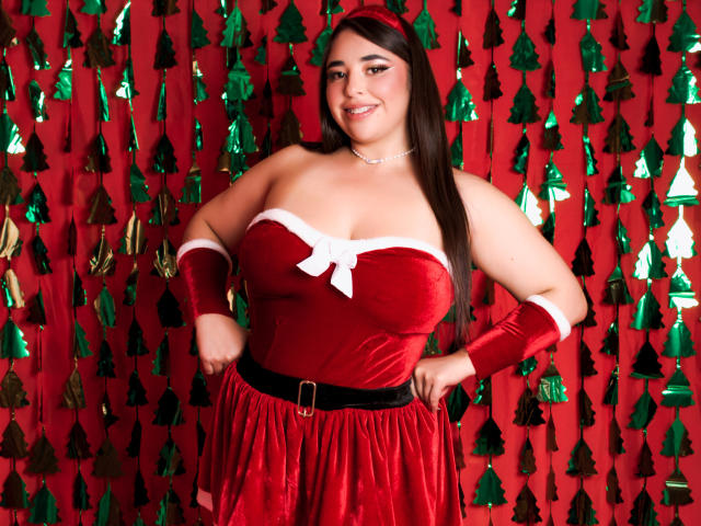 BaGiolett con grandes senos, vestida con un vestido navideño de terciopelo rojo con detalles blancos, un cinturón negro y guantes a juego, de pie con las manos en las caderas, sonriendo. Fondo rojo y verde..