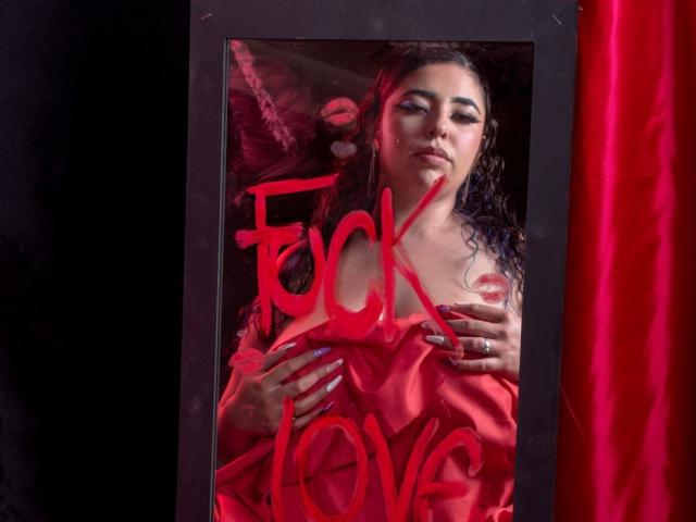 BaGiolett with curly dark hair, red lipstick, and blue nails, wearing an off-shoulder red dress, holding her tits, looking seductively at the camera, framed in black.