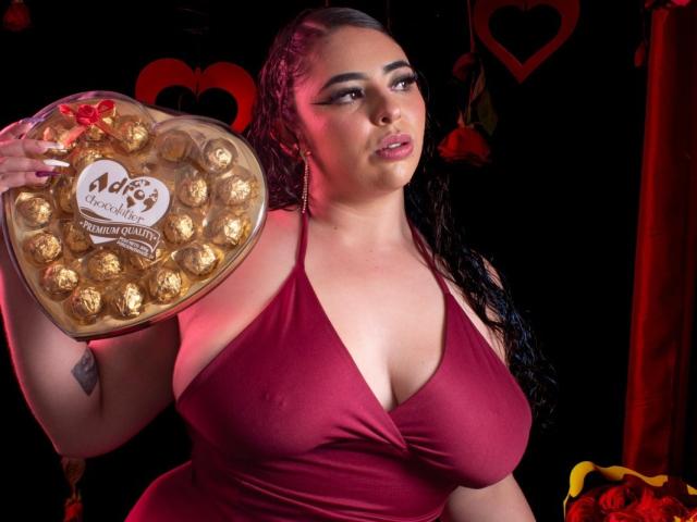 BaGiolett with big tits, wearing a red halter dress, holding a heart-shaped box of chocolate truffles, has long black hair, and a tattoo on her right arm.