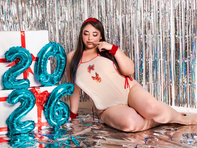 BaGiolett mit großem Hintern, trägt einen eng anliegenden, beigefarbenen Einteiler mit roten Akzenten und sitzt auf silberfoliertem Papier, neben blauen "2021"-Ballons und einer roten Geschenkbox..