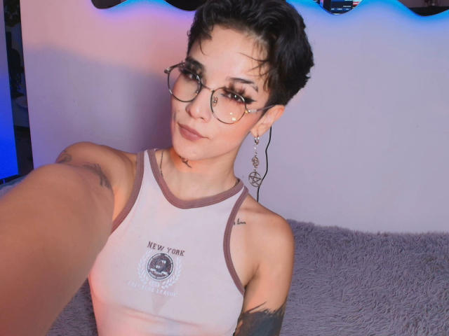 GiaMoretii with short black hair, glasses, and tattoos, wearing a white tank top, showing her tits, sitting on a gray fluffy couch, smiling at the camera.