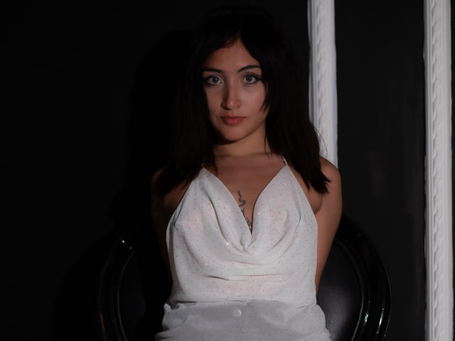 KaylovRyder with dark hair, wearing a white, halter-neck dress, showing small tits, sitting in a black chair, looking at the camera with a serious expression.
