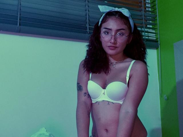 KaylovRyder with wavy brown hair, glasses, white bow, yellow bra, and no panties, showing small tattoos on her right arm, standing in a green-lit room.