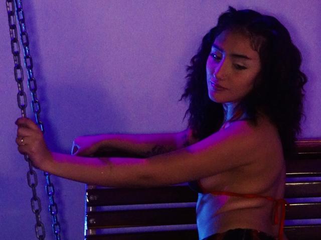KaylovRyder with curly hair, holding a chain, wearing a red string bikini top, exposing her small tits, and a tattoo on her arm, in blue-purple lighting.
