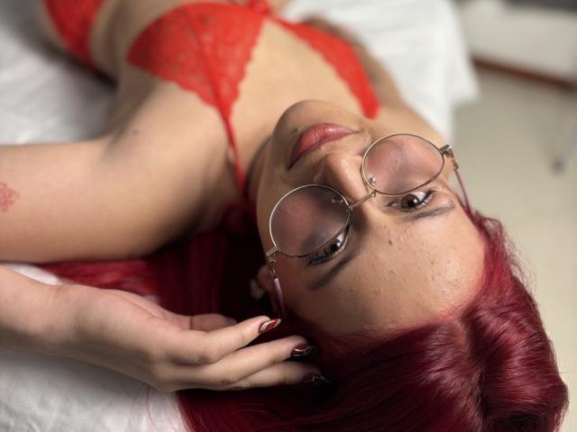 CherryMist with red hair, glasses, and red lingerie, lying on a bed, looking up, hand on her head, showing her tits, with a slightly flushed face.