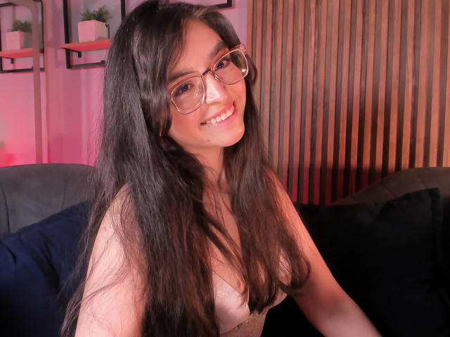 EvaMcAllister with long black hair, glasses, smiling, showing her small tits, wearing a low-cut top, sitting on a black couch, wooden wall in the background.