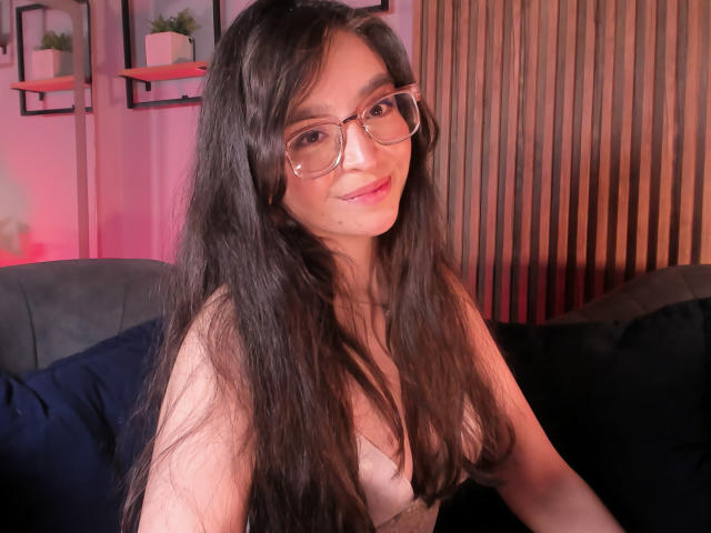 EvaMcAllister with long black hair, glasses, and pink lipstick, sitting on a dark couch, smiling, showing her small tits in a pink-lit room.