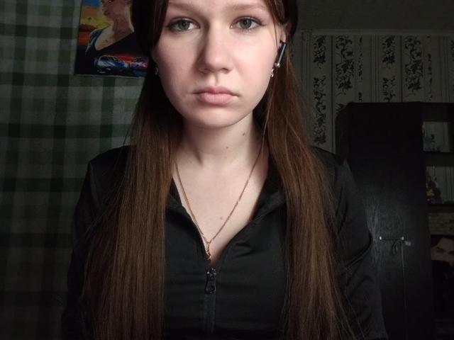 Kary Whitler, met lang bruin haar, bleke huid, gekleed in een zwart shirt met rits, met een serieuze blik, heeft kleine borsten en een dunne ketting, in een slecht verlichte kamer..