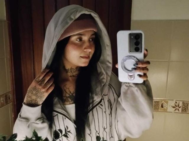 GabyMathew with tattoos, wearing a hoodie and pinkish headscarf, taking a mirror selfie with a white phone, showing off her tits and tattoos.