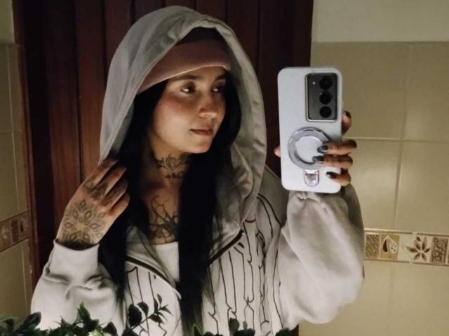 GabyMathew with tattoos, wearing a hooded sweatshirt and pinkish headscarf, holding a phone with a ring, taking a mirror selfie, showing dark hair and a subtle smile.