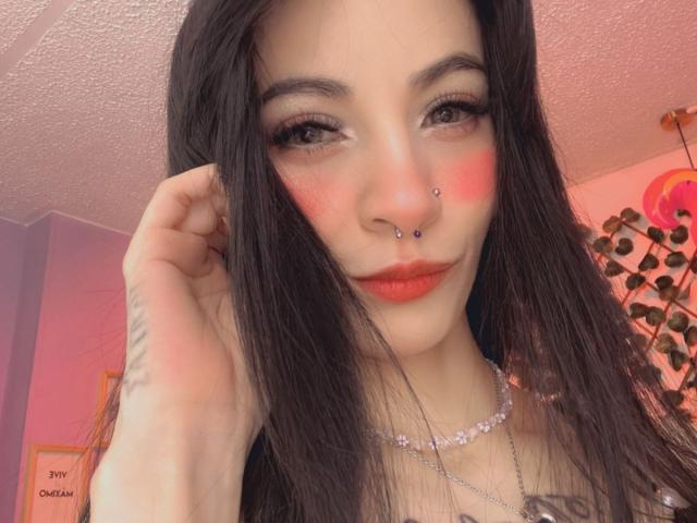 LaurenEvie69 with black hair, red blush, nose piercings, and red lipstick, looking at the camera with a slight smile. She's wearing a necklace and touching her hair.
