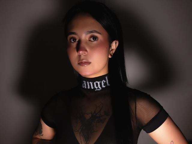 GabyMathew with black hair, sheer black top, visible chest tattoo, earrings, and "angel" collar, looking at camera with neutral expression. Soft lighting.