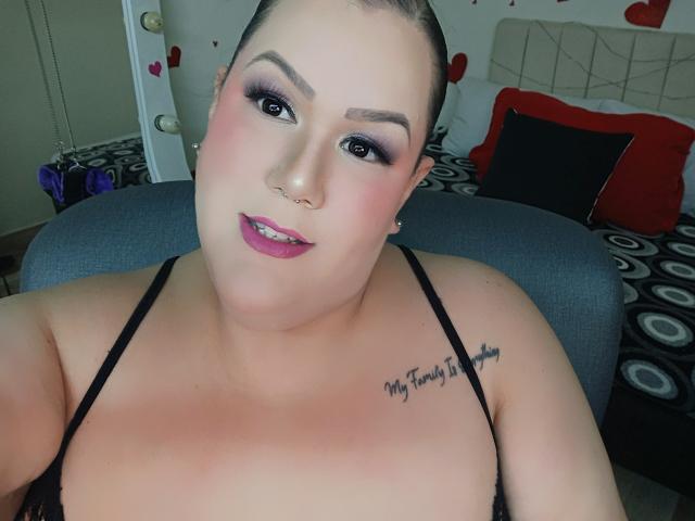 GiiannaMorrys has pale skin, dark hair in a bun, purple lipstick, and a tattoo on her right shoulder. She's wearing a black strap top, showing her boobs.