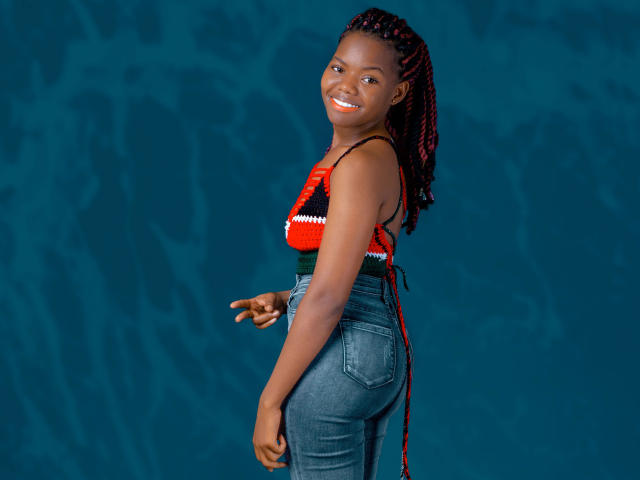 DottieSweet, à la peau foncée, avec des cheveux tressés noirs et rouges, souriante, portant un haut tricoté rouge et noir et un jean bleu taille haute, pointant de la main droite..