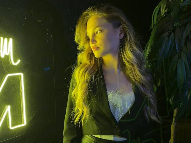 JamilaRomeiro with long, wavy blonde hair in a black and green jacket, white lace top, looking to the side, illuminated by yellow-green light, holding a plate.