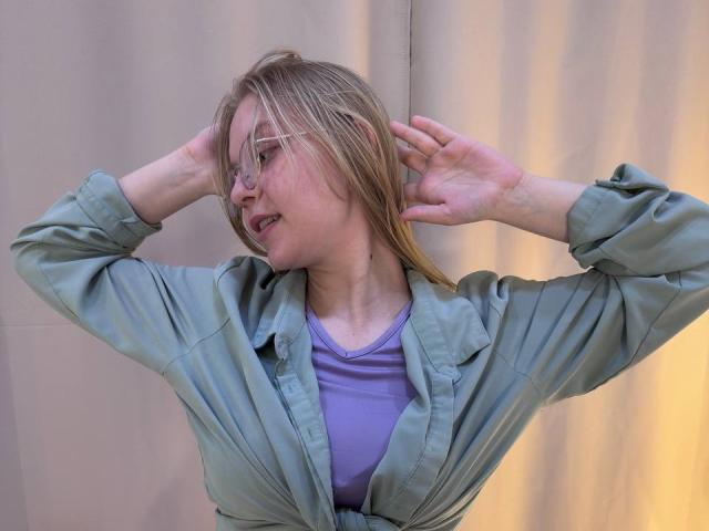 QueenBooser, met blond haar, draagt een groene blouse en een paars top, heeft haar armen omhoog, haar ogen zijn gesloten en ze heeft een lichte glimlach, voor een beige gordijn..