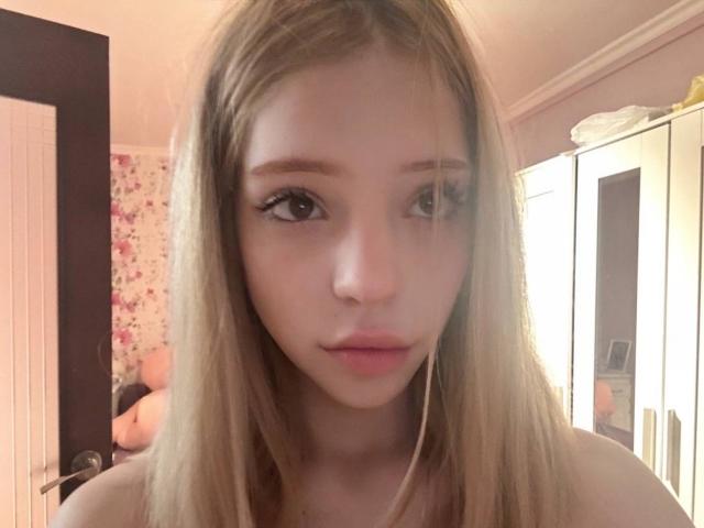 SiobhanSvancara has long blonde hair, brown eyes, and a serious expression with slightly parted pink lips, looking straight at the camera in a softly lit room.