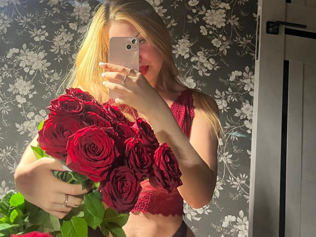 NicoleHrum with blonde hair, red lace top, holding a bouquet of red roses, taking a mirror selfie with a rose gold iPhone, her ass slightly visible in the reflection.