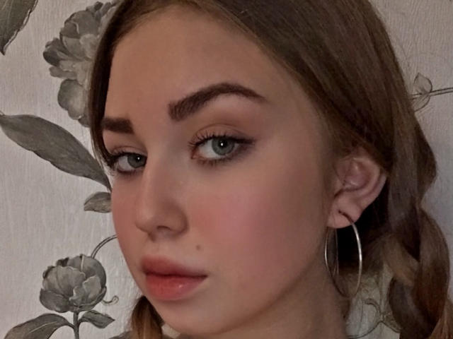 NicoleHrum has blue eyes, brown eyebrows, pink lips, and a small moles' on her left cheek. She's wearing a silver hoop earring in her brown braided hair.