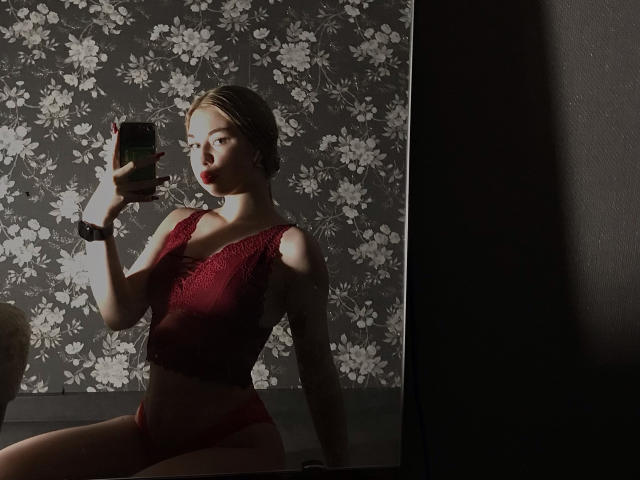 NicoleHrum with light skin, blonde hair, red lace bra, and matching red panties, holding a phone, taking a mirror selfie, showing her tits and ass in dim light.