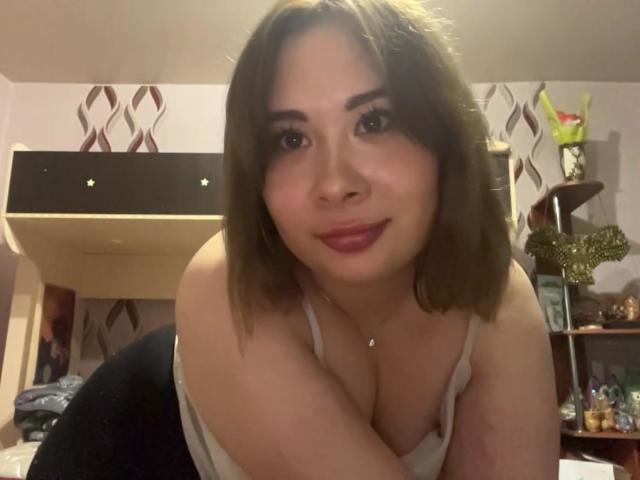 JoshuaShor, com cabelo castanho-claro, pele clara e batom vermelho, inclinada para frente, mostrando seios grandes em uma blusa branca, sorrindo suavemente, em um quarto com iluminação quente..