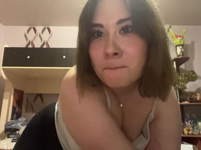 JoshuaShor, con cabello castaño hasta los hombros, un top blanco y senos visibles, inclinado hacia adelante, mirando a la cámara con una ligera sonrisa, en una habitación con poca luz..