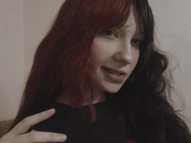 Jaditta, avec ses cheveux ondulés roux et noirs, son anneau au nez et son piercing à la lèvre inférieure, souriant discrètement, portant une chemise noire et touchant son épaule..