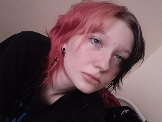 Jaditta, met roze en zwarte gekleurd haar, blauwe ogen, een neusring, een lip piercing en oorbel piercings, draagt een zwart shirt en kijkt iets opzij..