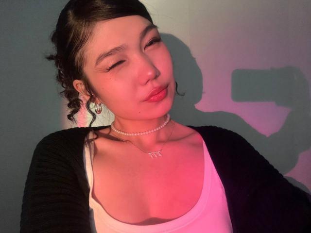 AmeliyaAyrton with closed eyes, pink lips, black cardigan, white tank top, pearl necklace, earrings, and braided hair, showing a shadow of a camera on a pinkish wall.