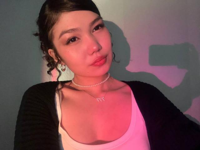 AmeliyaAyrton with pouty red lips, white tank top, black cardigan, pearl necklace, and earrings, has cute face and small tits, brown hair in a bun, in a selfie.