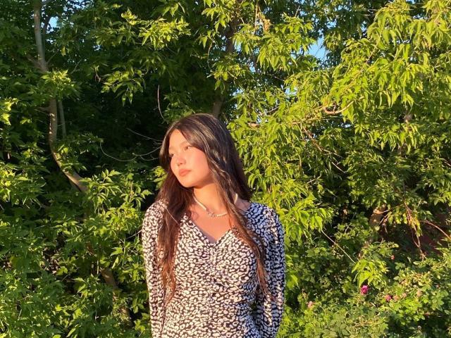 AmeliyaAyrton with long brown hair, wearing a patterned dress, standing in sunlight, green leaves in background, sunlight on her face, looking to the side, small pink flower.