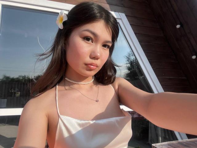 AmeliyaAyrton with long brown hair, white flower in hair, wearing white spaghetti dress, pearl choker, and necklace, taking selfie, showing small tits, light pink blush, sunny outdoor.