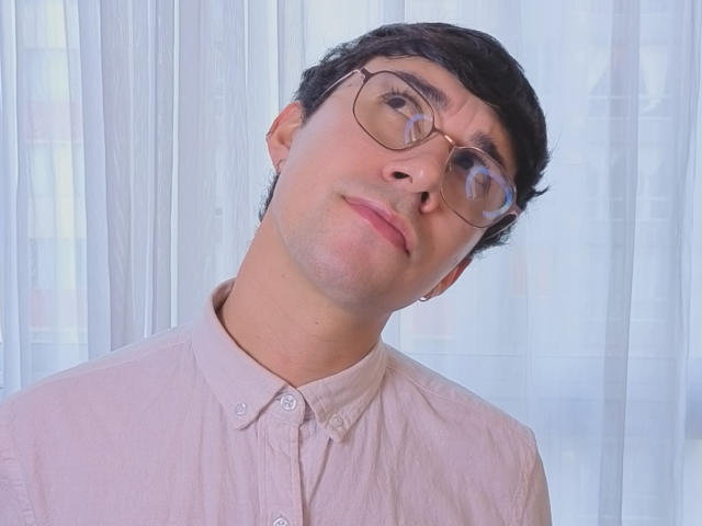 OliverHot69 with glasses, light pink shirt, and slightly tilted head, looking up with a subtle smile. Medium skin, black hair, and a small earring in his left ear.