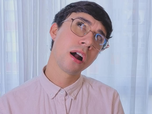 OliverHot69 with glasses, short dark hair, light pink shirt, mouth open in surprise, blue reflection in right eye, soft light, slightly tilted head.