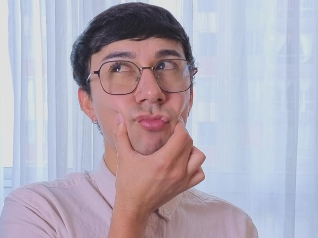 OliverHot69, with glasses and dark hair, puffs out cheeks, resting hand on chin. Wearing a pink shirt, his fingerless pinkie is raised. He's looking to the side.