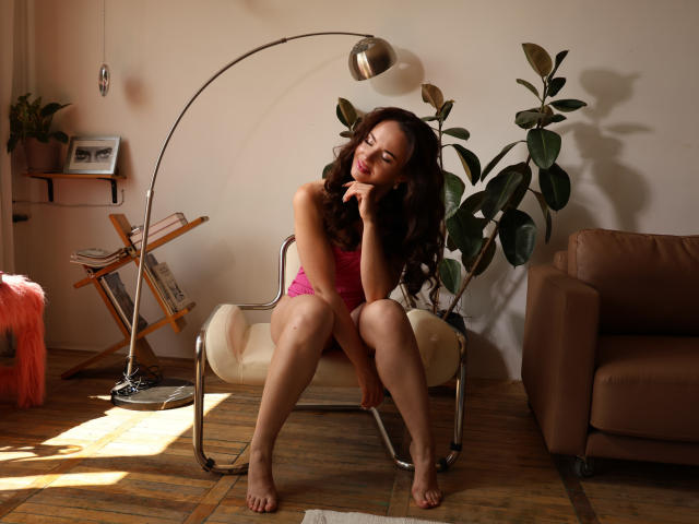 KaraCriegs with wavy brown hair, pink lingerie, sitting on white chair, legs apart, hand on chin, arched lamp, brown armchair, potted plant, wooden floor, sunny room.