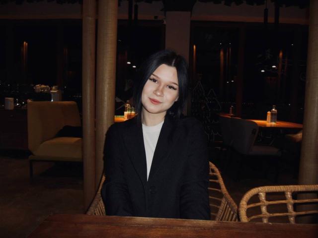 SaraJerome with black hair, pale skin, and red lipstick, wearing a black blazer over a white shirt, sitting at a wooden table in a dimly lit room.