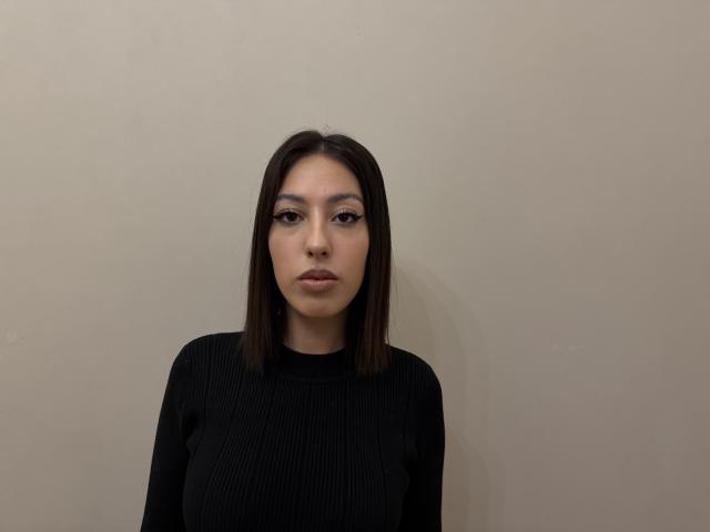 DeannDelcamp with medium brown hair, wearing a black ribbed long-sleeve top, has a neutral expression, looking directly at the camera, standing against a plain beige wall.