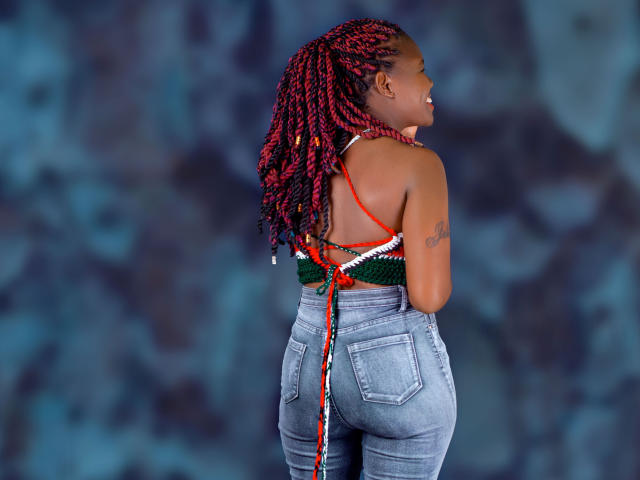 DottieSweet met rood en zwart gevlochten haar, een groen gebreid topje met rode bandjes, blauwe jeans, een tatoeage op haar rechterarm, lachend, met de rug naar de camera, waarbij haar achterwerk zichtbaar is..