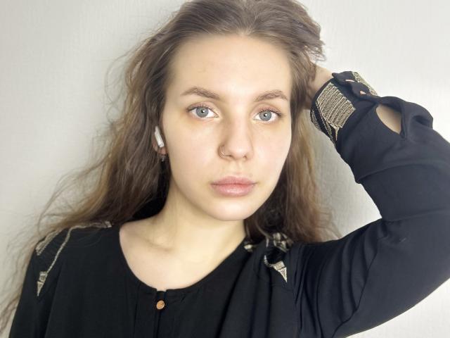 JamikaAdule with wavy brown hair, blue eyes, and a nose ring, wearing a black shirt, has a serious expression and her right arm raised, showing a wristwatch.