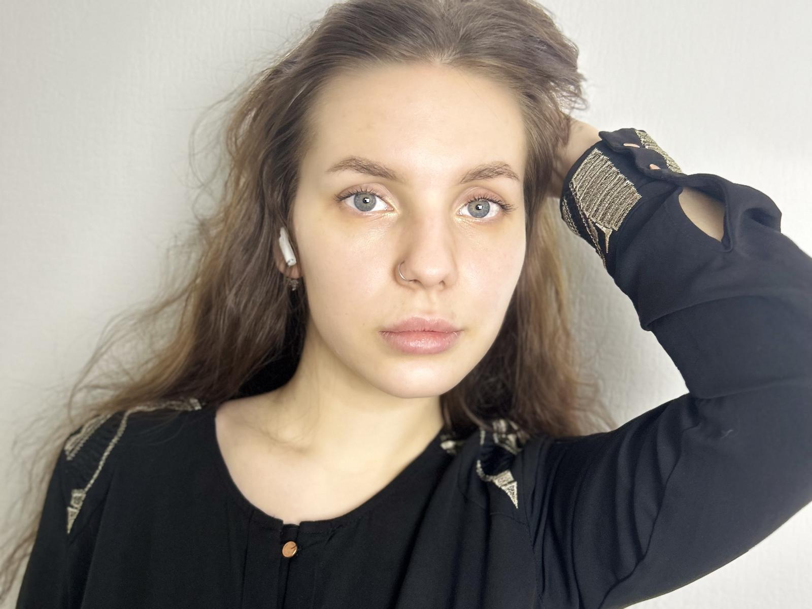Jamika Adule, met golvend bruin haar, blauwe ogen en een neusring, draagt een zwarte blouse en heeft een serieuze uitdrukking. Haar rechterarm is omhoog, waarbij ze een polshorloge laat zien..