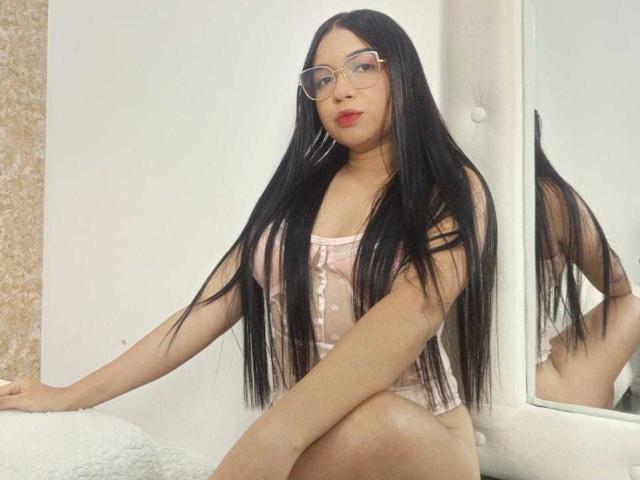 AliceSmity with long black hair, glasses, and pink polka-dot lingerie, sitting, showing her ass, small tits, and red lips, in a mirror-reflected room.