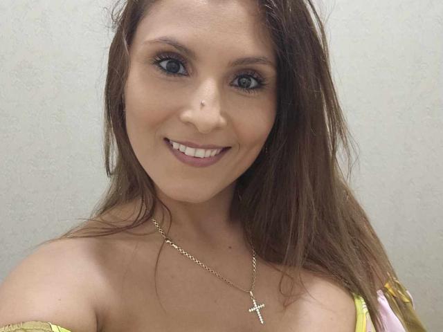 DianeExo, com cabelo castanho, olhos azuis, sorrindo, usando um colar com uma cruz dourada, blusa amarela com um ombro descoberto, mostrando seus seios, contra um fundo simples..