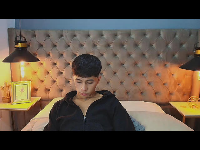 SaintDangelo with short black hair, wearing a black zip-up hoodie, sits on a bed with a tufted, beige headboard, looking down, flanked by black hanging lights.