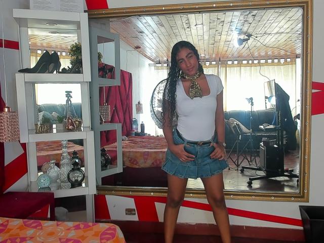 NatalyBlonder with long black hair, white tight shirt, blue denim skirt, standing in front of a mirror, hands on skirt, necklace, smiling, mirror reflecting room, camera on right.